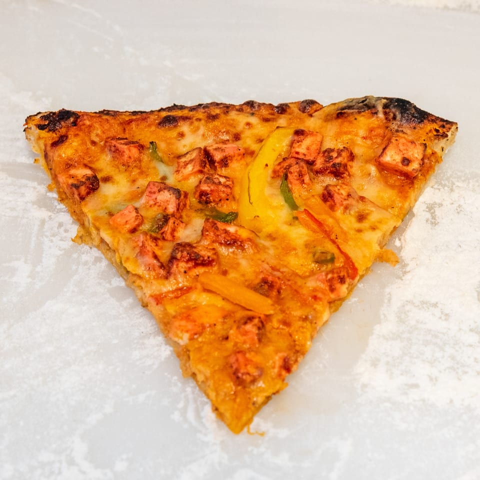 Triangular slice of cheesy pizza topped with diced vegetables and browned edges on white paper.