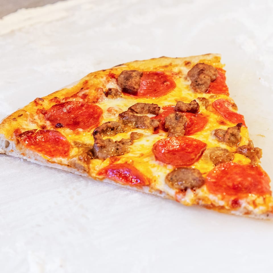 Triangular slice of cheese pizza topped with pepperoni and sausage on white paper.