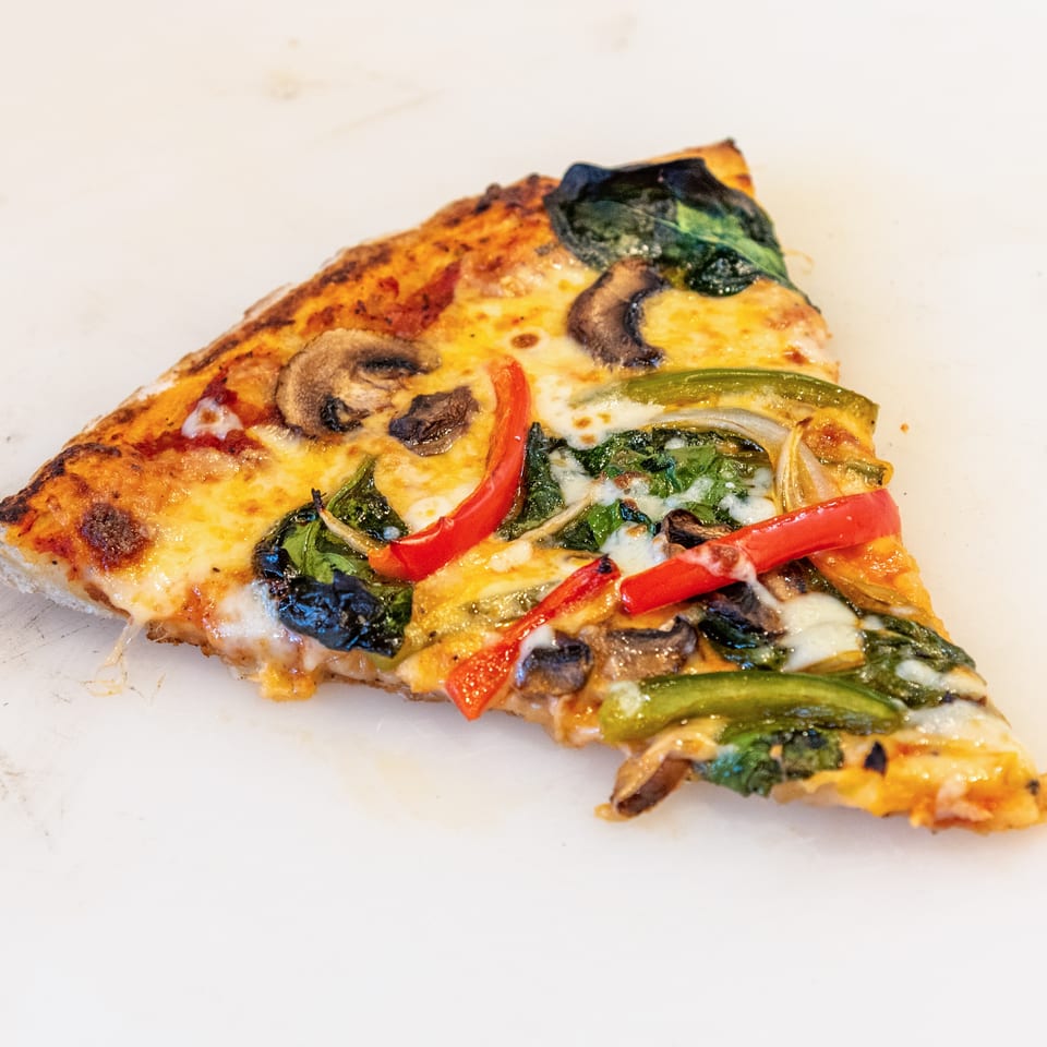 Slice of cheesy vegetable pizza topped with mushrooms, spinach, and red and green peppers on a white surface.